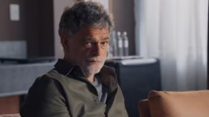 A foto mostra o ator Eduardo Moscovis que interpreta o Rogério. Ele é um homem branca, com barba e cabelo grisalhos, usando uma camisa jeans cinza.
