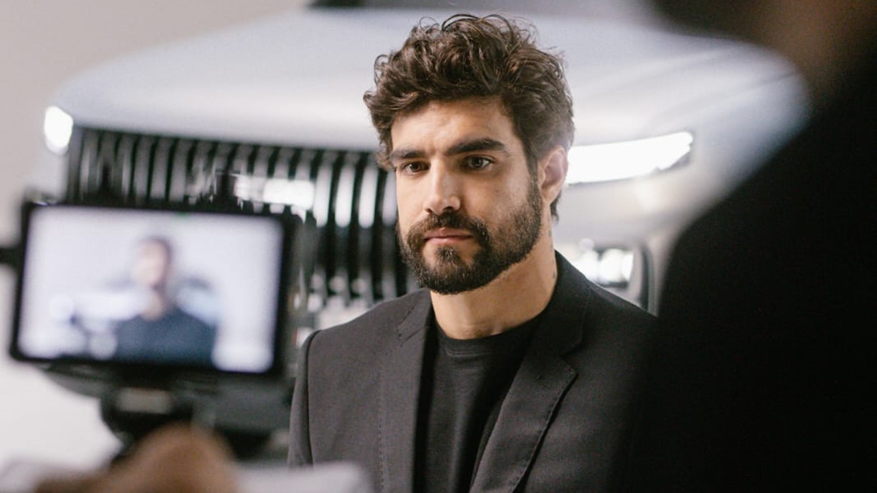 A imagem mostra o Caio Castro. Um homem branco, de cabelo castanho curto e com barba, usando terno.