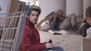 Homem branco, com barba, sujo e sentado na calçada em situação de rua.