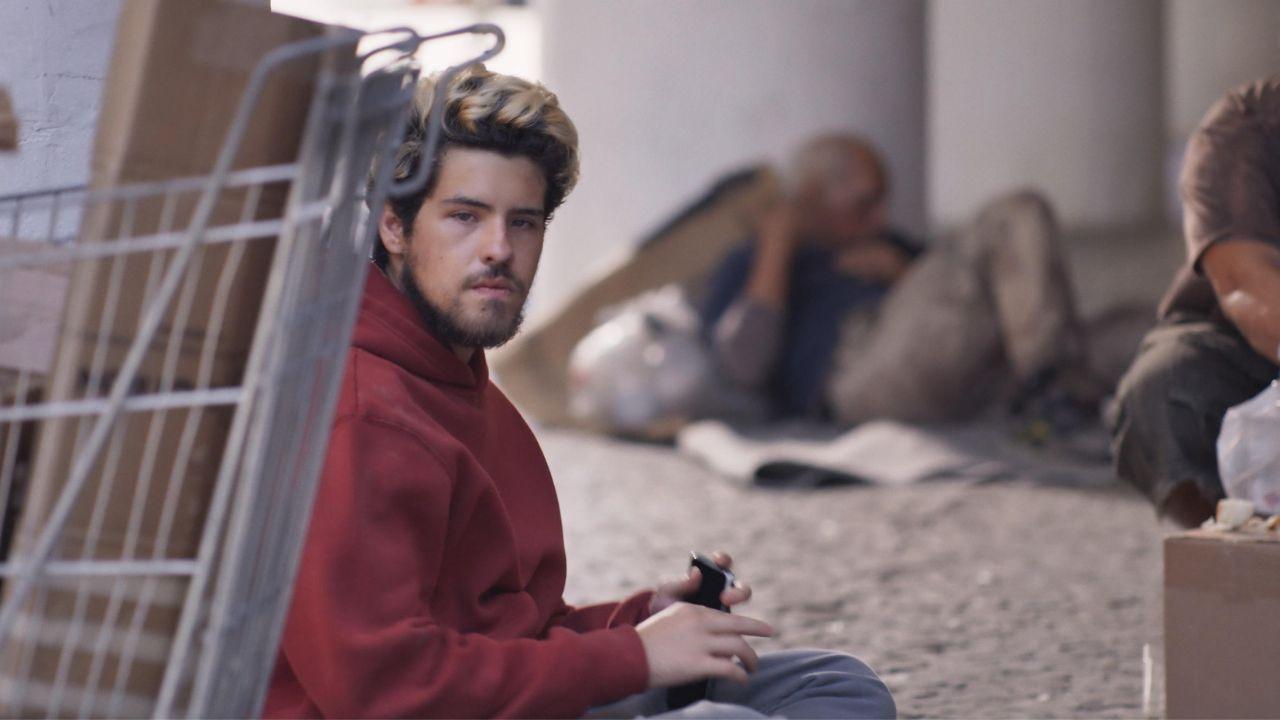 Homem branco, com barba, sujo e sentado na calçada em situação de rua.