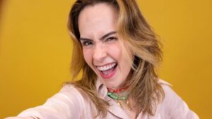 Mulher branca, loira, cabelo ondulado, sorrindo para a câmera e segurando o celular na selfie.