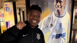 Homem negro, com moletom preto com o símbolo do Corinthians. Ele está do lado de uma foto dele em tamanho real quando era mais novo e jogava futebol.