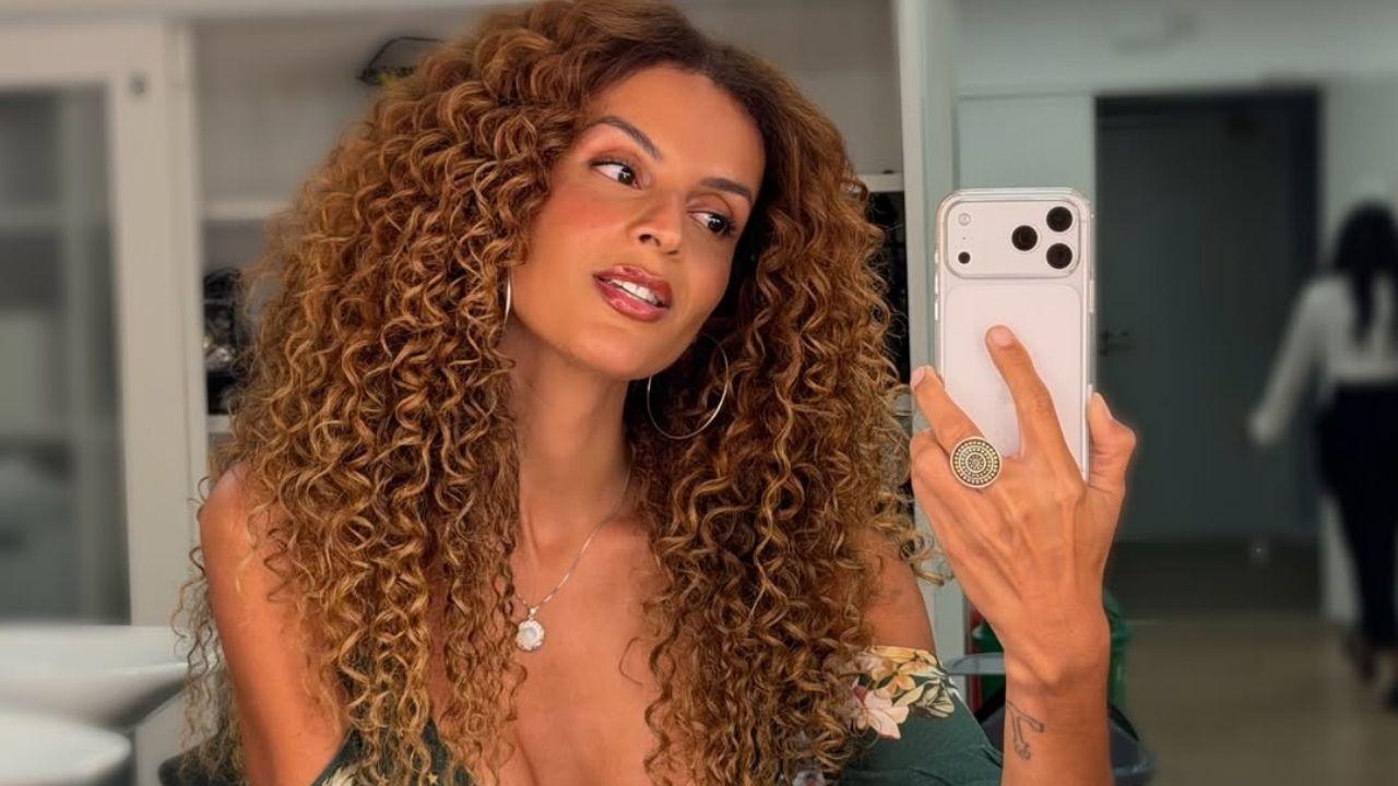 Mulher negra, cabelo cacheado cheio, segurando um celular, tirando foto no espelho.