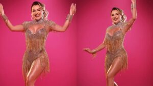 Mulher branca, loira, com fantasia de carnaval de correntes e glitter em um fundo rosa.
