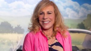 Mulher branca, idosa, loira, usando camisa rosa e sorrindo.