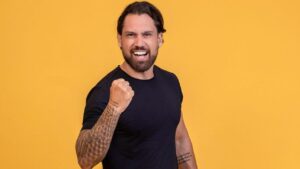 Homem branco, cabelo castanho, barba, camiseta preta em um fundo amarelo.