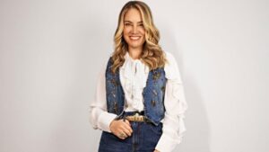 Juliana Baroni. Mulher branca, loira, cabelo ondulado, usando camiseta branca com colete jeans.