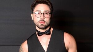 Rico Melquiades. Homem branco, cabelo preto curto, de óculos, colete e gravata preto.