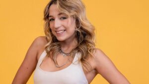 Mulher branca, loira, usando saia curta e blusa branca num fundo amarelo.