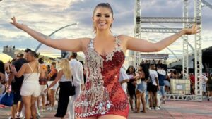 Mulher branca, cabelo castanho preso em um rabo de cavalo. Ela usa um vestido de paetê vermelho curto. Ao fundo tem pessoas ao ar livre.