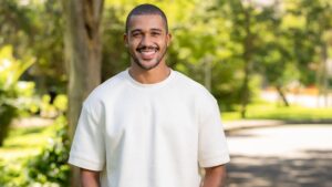 Homem negro, careca, com barba, usando camiseta branca. No fundo, tem árvores desfocada na foto.