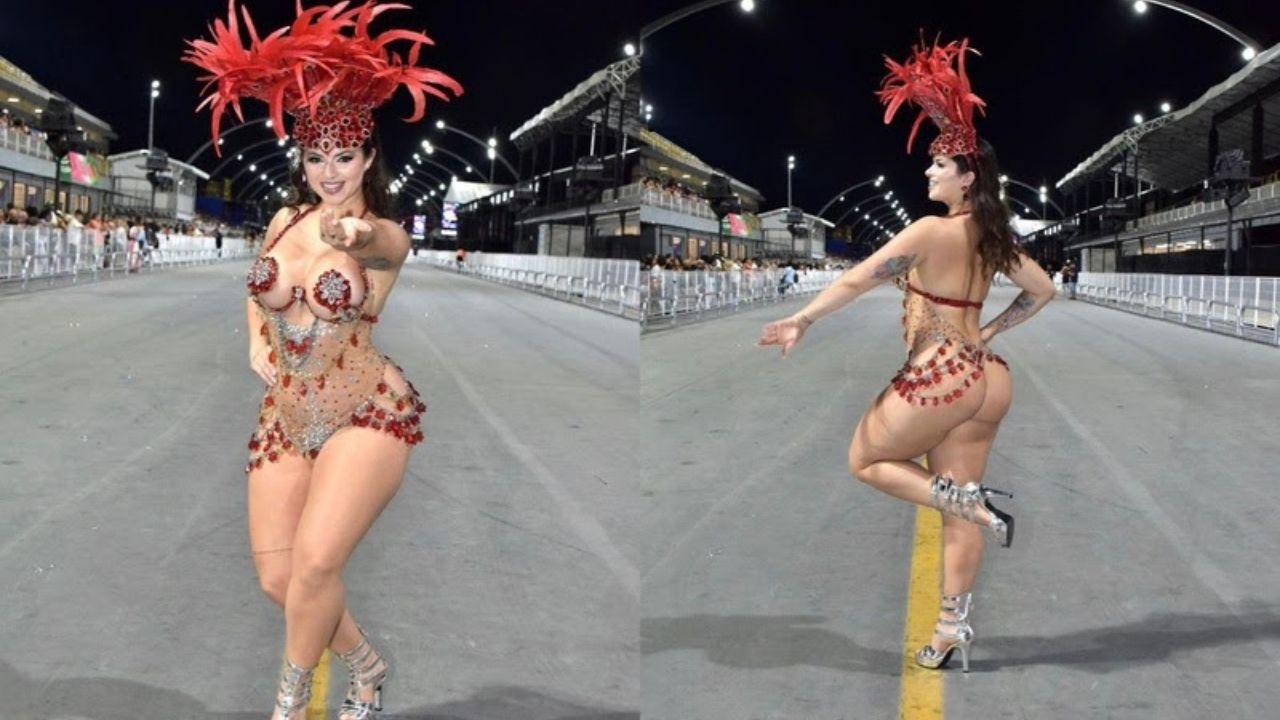 Mulher branca, cabelo castanho preso com fantasia de carnaval vermelha na avenida. É uma fantasia cheia de strass e curta com penas na cabeça.
