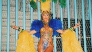Sabrina Sato. Ela está encostada na parede com fantasia de carnaval cheia de plumas na cabeça, strass e brilho no corpo. As cores predominantes são amarelo e verde.