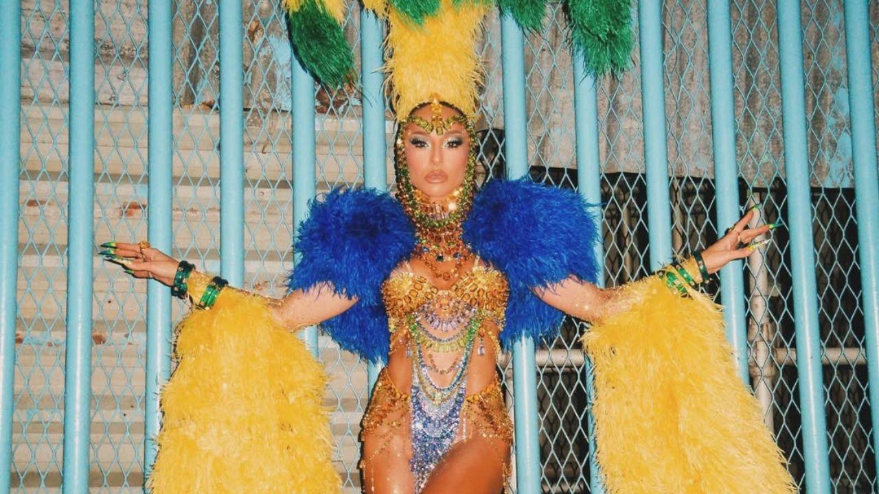 Sabrina Sato. Ela está encostada na parede com fantasia de carnaval cheia de plumas na cabeça, strass e brilho no corpo. As cores predominantes são amarelo e verde.
