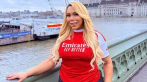 Mulher branca, cabelo loiro comprido, usando camiseta de time vermelha. Ao fundo, prédios, rio e uma roda gigante.