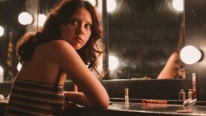 Mia Goth. Mulher branca, cabelo castanho preso, sentada num camarim.