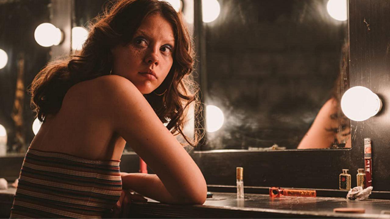 Mia Goth. Mulher branca, cabelo castanho preso, sentada num camarim.