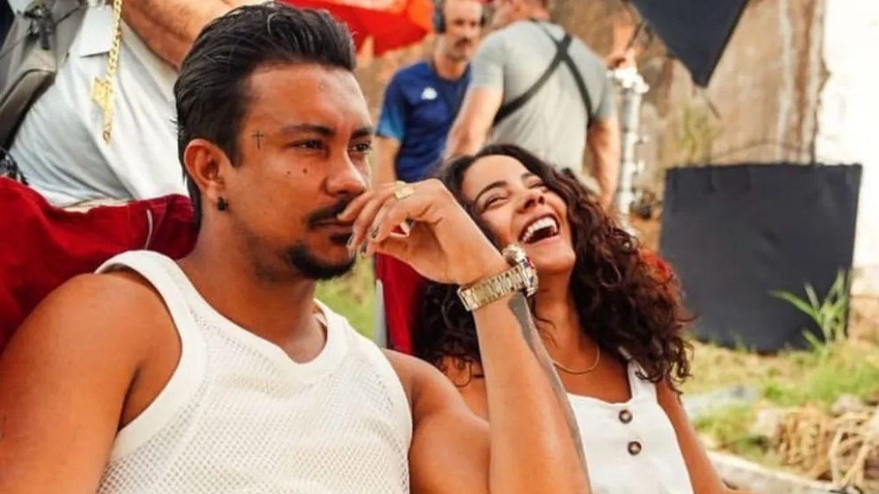 Xamã e Bella Campos sentados ao ar livre enquanto ele faz cara de sério e ela dá gargalhadas.