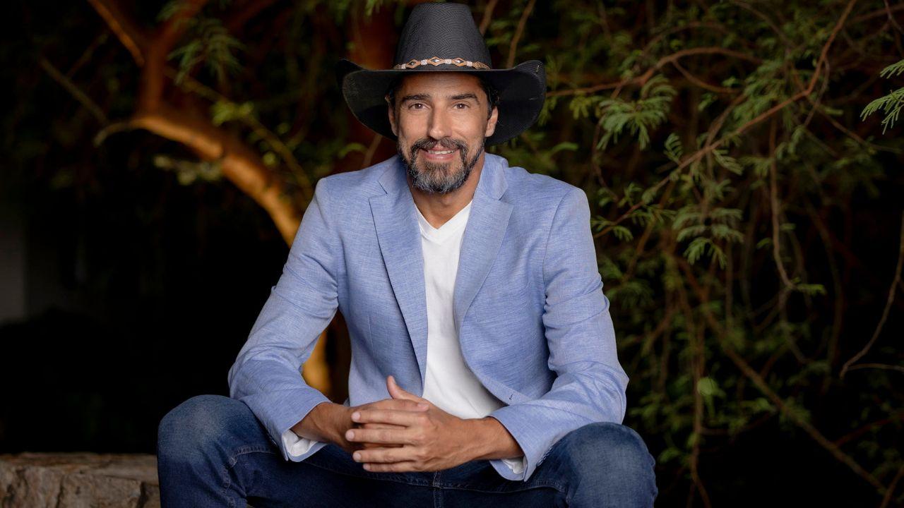 Alberto Cowboy. Homem branco, barba e cabelo castanho, usando chapéu, sentado no banco, usando terno azul e sorrindo.