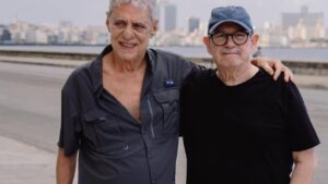 Chico Buarque ao lado de um amigo. Ambos homens idosos e brancos, em pé ao ar livre.