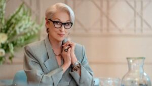 A atriz Meryl Streep de Miranda Priestly no filme O Diabo Veste Prada. Ela está sentada numa mesa mostrando os clássicos cabelos brancos em corte curto.