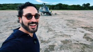 Lucas Estevam. Homem branco, cabelo preto curto, usando óculos de sol, camiseta azul e na frente de um helicóptero ao ar livre.