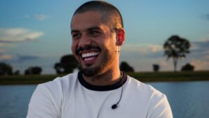 Zé Felipe com cabelo raspado, sorrindo, camiseta branca, ao ar livre.