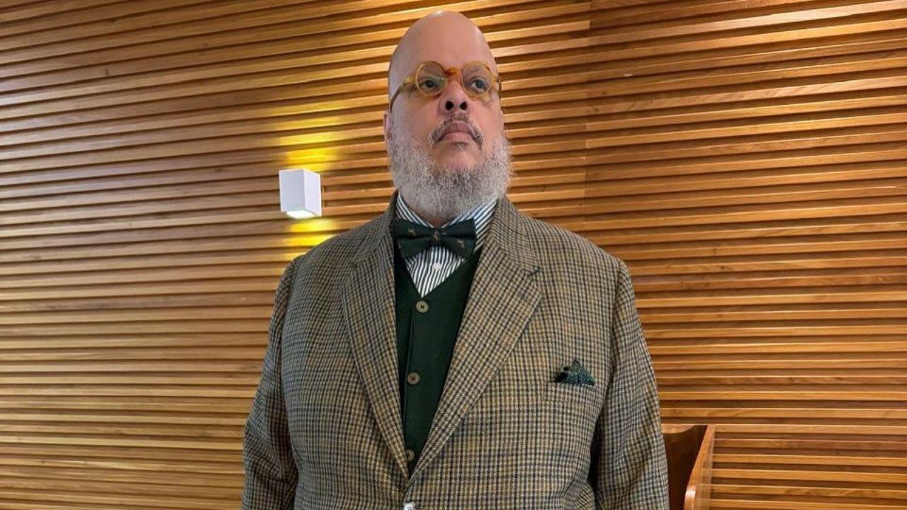 Foto mostra o cantor Ed Motta. Homem negro, usando terno marrom claro, óculos e careca.