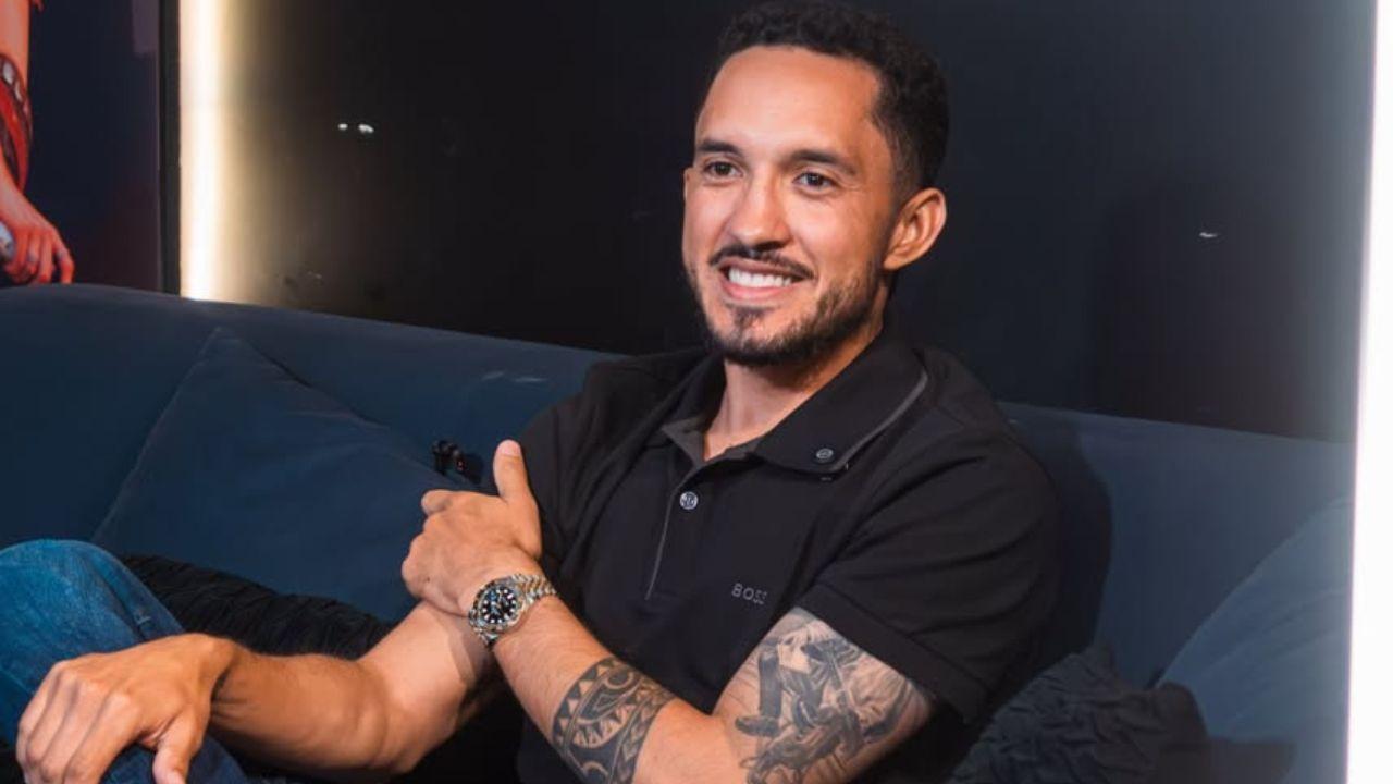 Cantor Grelo sentado numa poltrona em entrevista. Homem branco, cabelo castanho, usando camiseta preta.