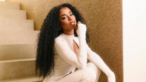Influenciadora Lorena Maria. Mulher negra, cabelo cacheado longo, usando vestido branco agachada na escada.