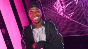 Vini Jr. Homem negro, usando boné preto, moletom preto e sorrindo.