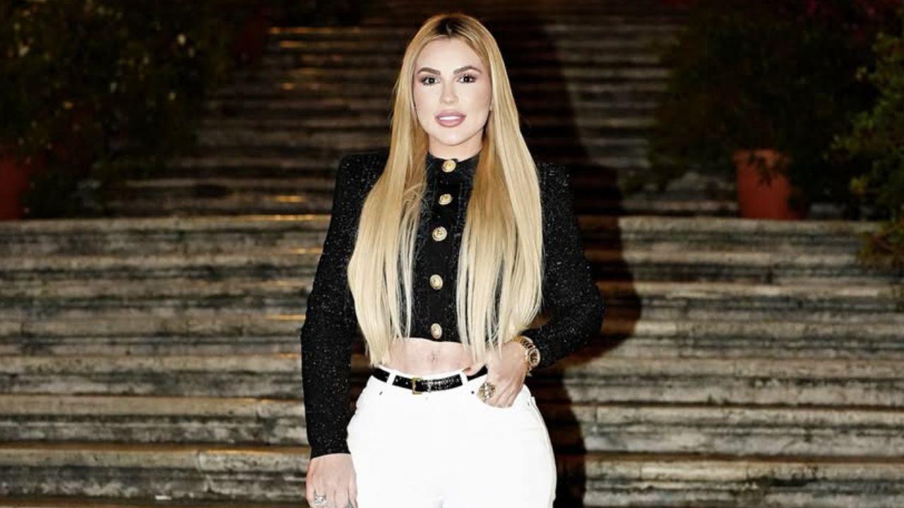 Influenciadora Deolane Bezerra. Mulher branca, loira, cabelo longo liso, com blusa preta e calça branca.