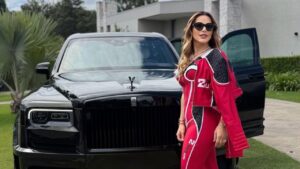 Deolane com macacão vermelho, usando óculos de sol, na frente de um carro de luxo no quintal da sua mansão.