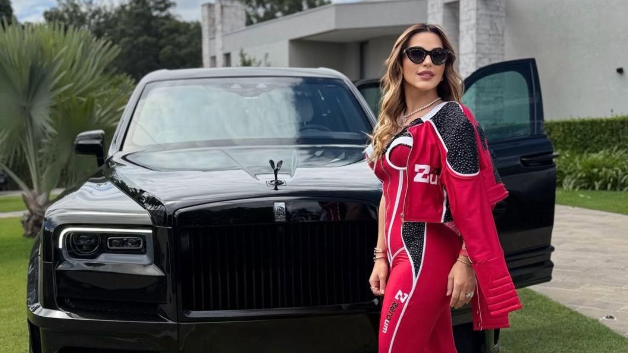 Deolane com macacão vermelho, usando óculos de sol, na frente de um carro de luxo no quintal da sua mansão.