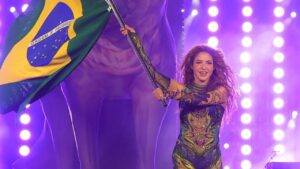 Shakira com collant roxo brilhante, segurando a bandeira do Brasil em show.