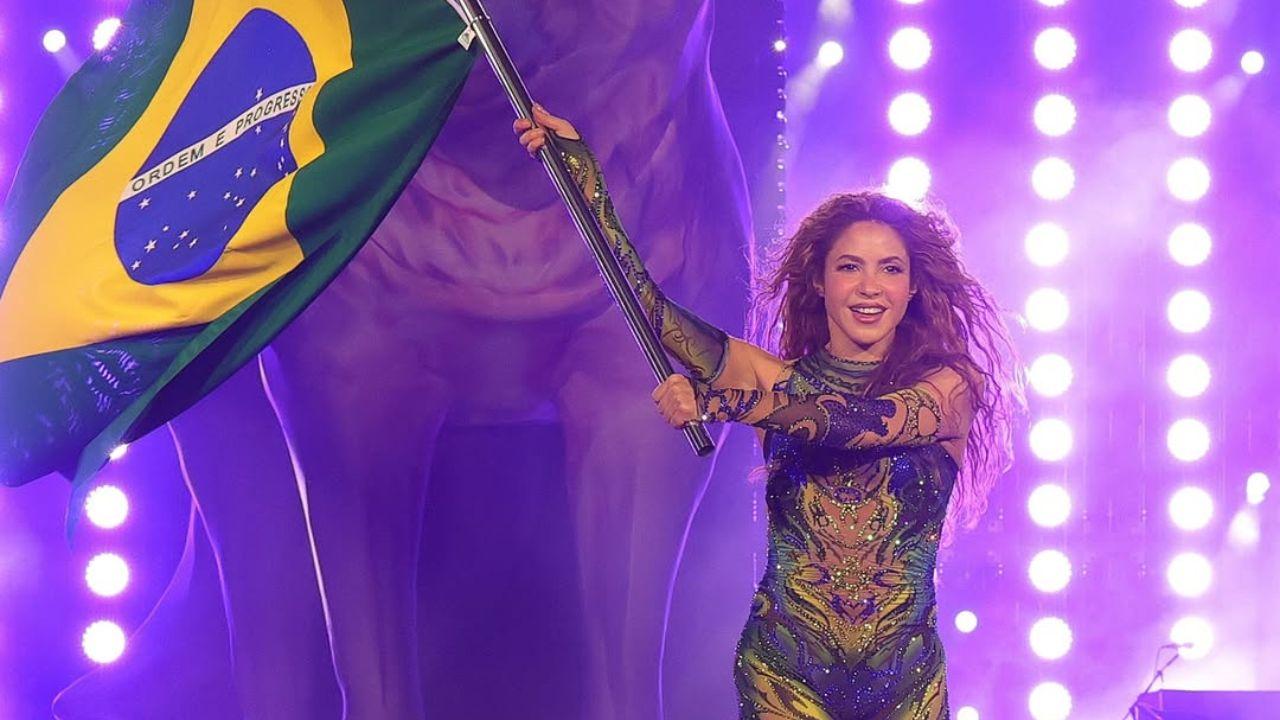 Shakira com collant roxo brilhante, segurando a bandeira do Brasil em show.