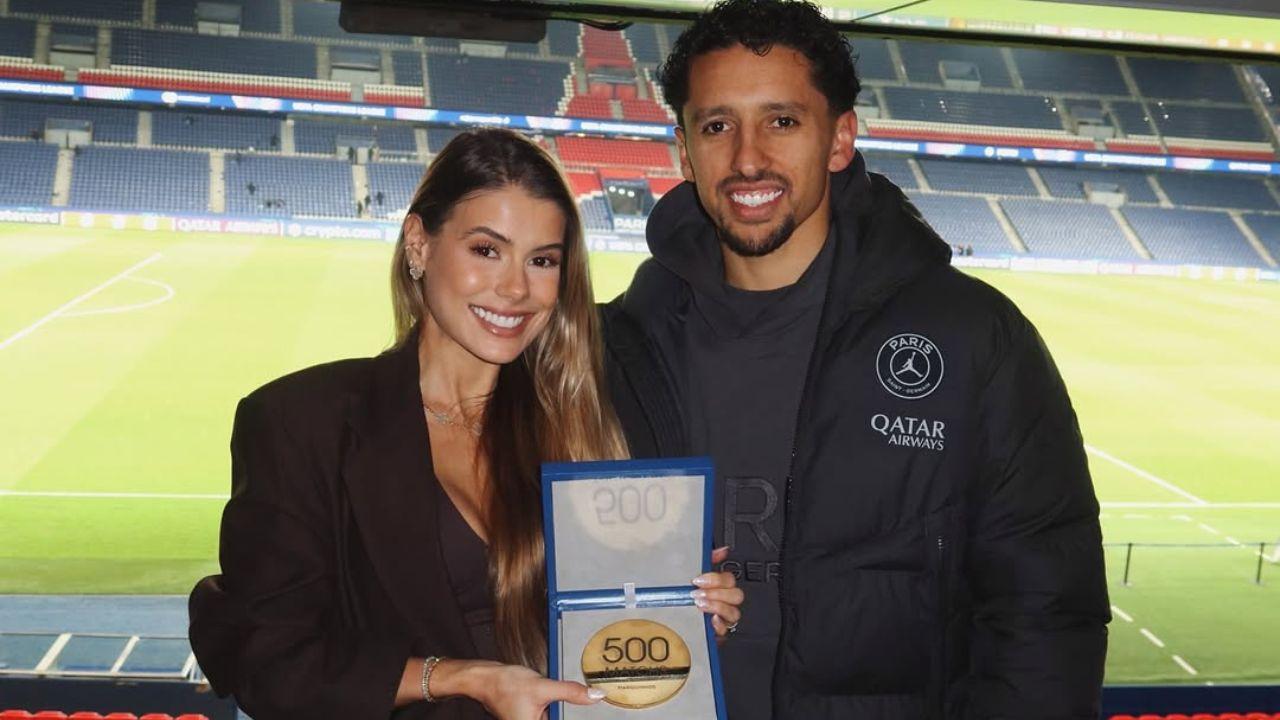 Carol Cabrino e Marquinhos no estádio de futebol. Mulher branca, cabelo loiro liso, homem negro, cabelo cacheado curto.