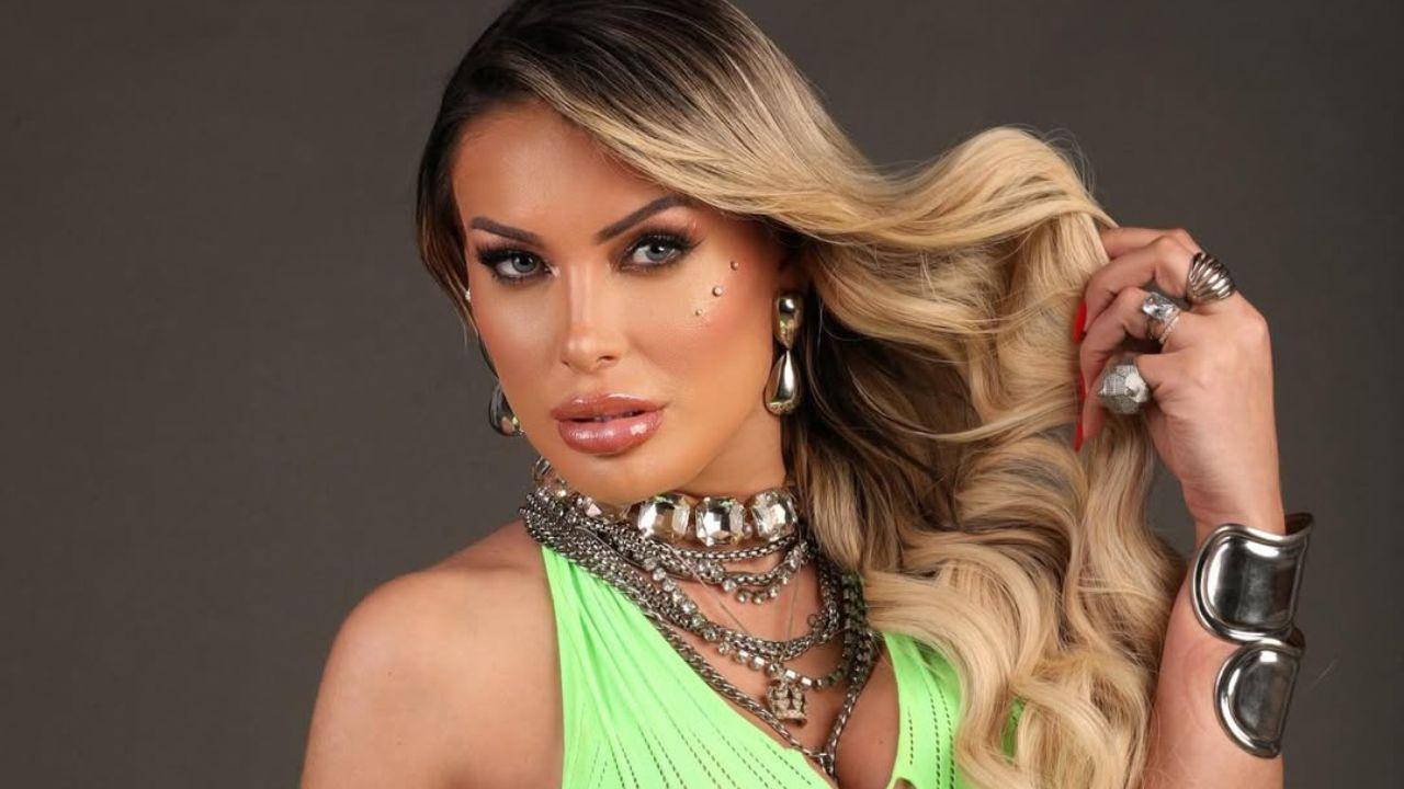 Foto mostra Andressa Urach. Mulher loira, bem maquiada, usando blusa verde e joias douradas.