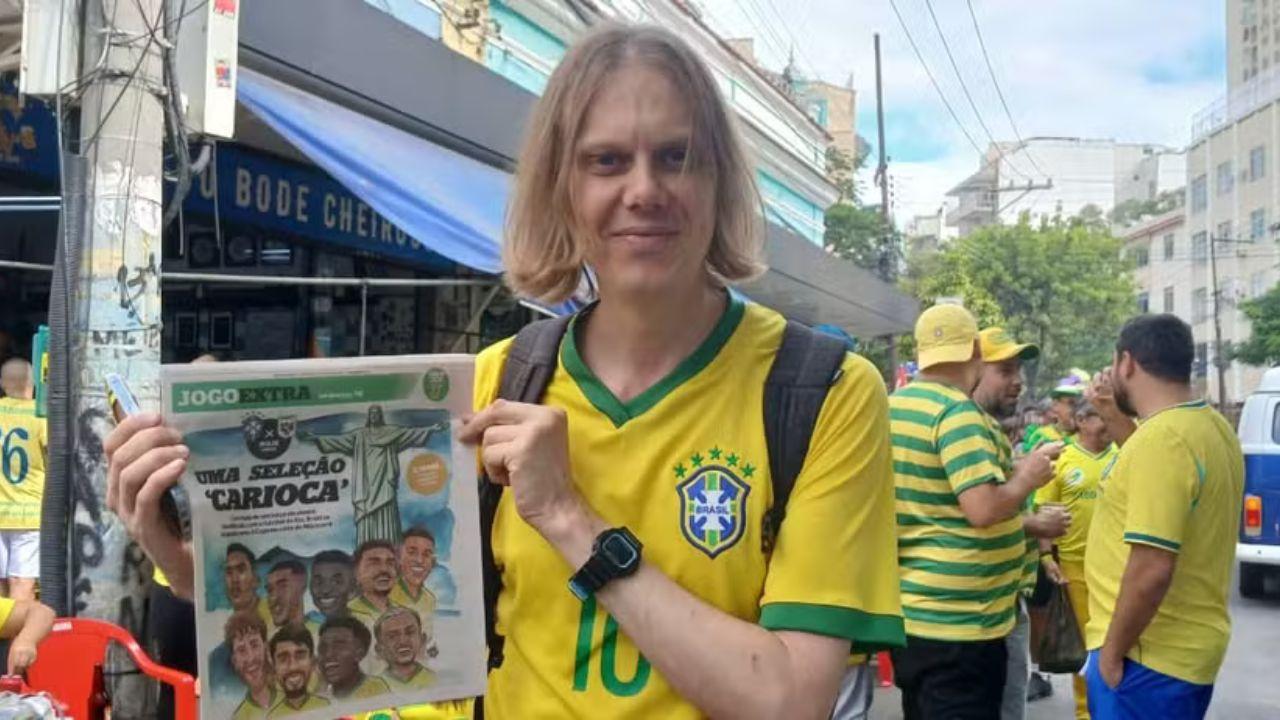 Foto mostra o torcedor Yuri Torsky no Brasil segurando um jornal do Extra. Ele é um homem branco, cabelo loiro liso com franja e usa a camiseta amarela da seleção brasileira.