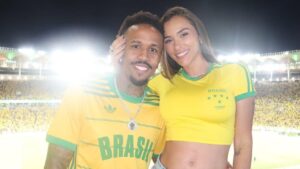 Tainá e Éder Militão usando camisetas do Brasil no estádio de futebol.