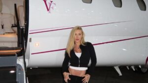 Foto mostra Virginia na frente do seu jatinho particular. Mulher branca, loira, cabelo liso, usando roupas esportivas pretas.