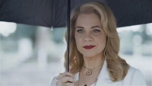 Foto mostra a atriz Isabel Teixeira interpretando a Pilar. Mulher branca, cabelo loiro, batom vermelho, ela está em um cemitério com um guarda-chuva.