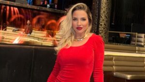 Clara Aguilar. Mulher branca, cabelo loiro, usando vestido e batom vermelho dentro de um cassino.