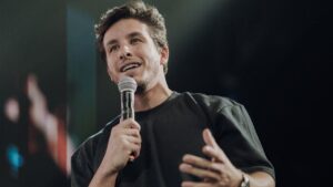 Foto mostra Ricky Tavares. Homem branco, cabelo castanho curto, usando camiseta preta, com um microfone na mão, falando no palco da igreja.