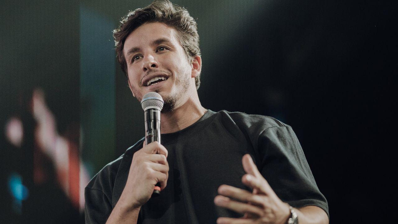 Foto mostra Ricky Tavares. Homem branco, cabelo castanho curto, usando camiseta preta, com um microfone na mão, falando no palco da igreja.