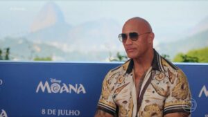 Foto mostra o ator Dwanye Johnson sentado, vestido de camisa estampada e óculos escuros.