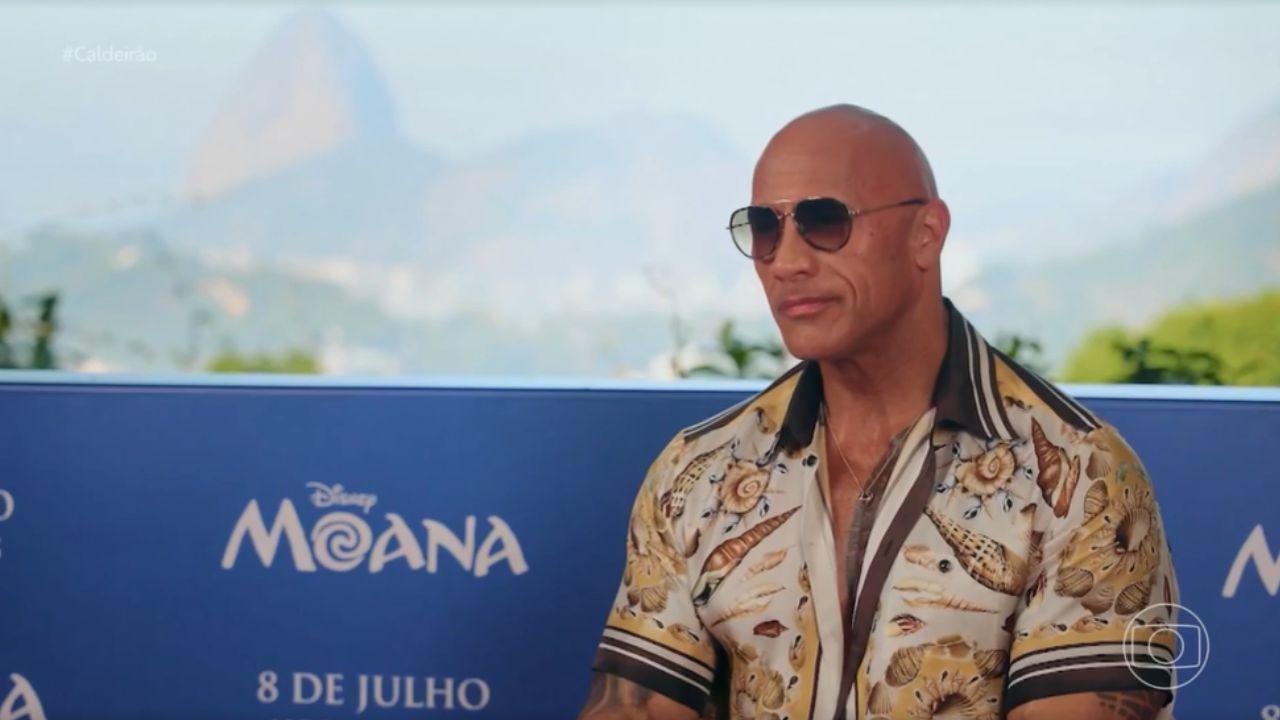 Foto mostra o ator Dwanye Johnson sentado, vestido de camisa estampada e óculos escuros.