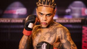 MC Cabelinho. Homem negro, de trança nagô, sem camisa, cheio de tatuagens, com luva de mma de luta.